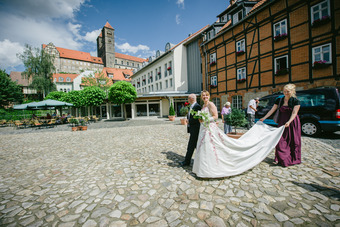 Braut im weißen Kleid mit Begleitung im Hof mit Kopfsteinpflaster, historischem Gebäude und Gästen im Hintergrund.