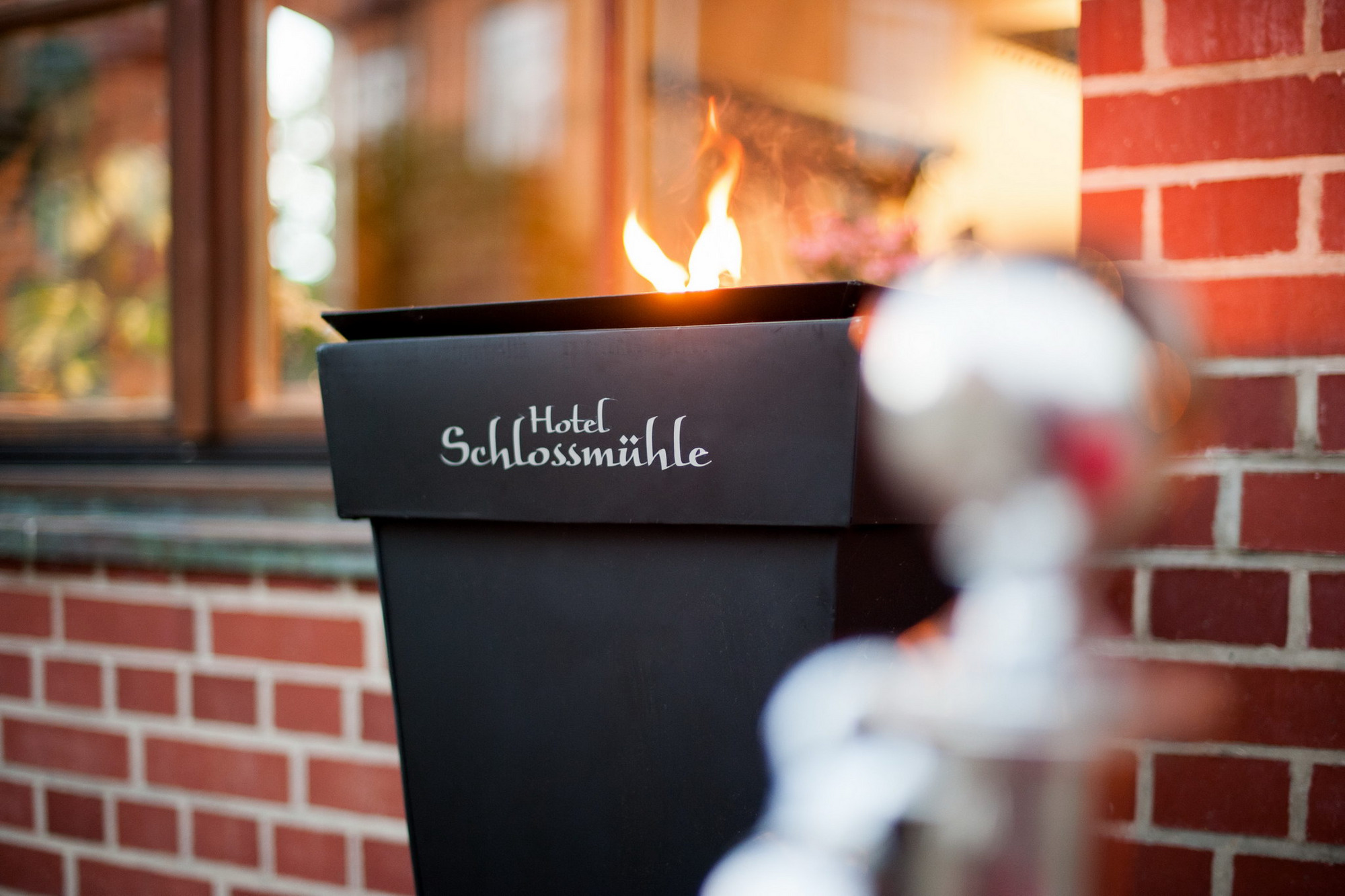 Schwarze Metallskulptur mit Flamme und Schriftzug „Hotel Schlossmühle“ vor Backsteinwand und Fenster.