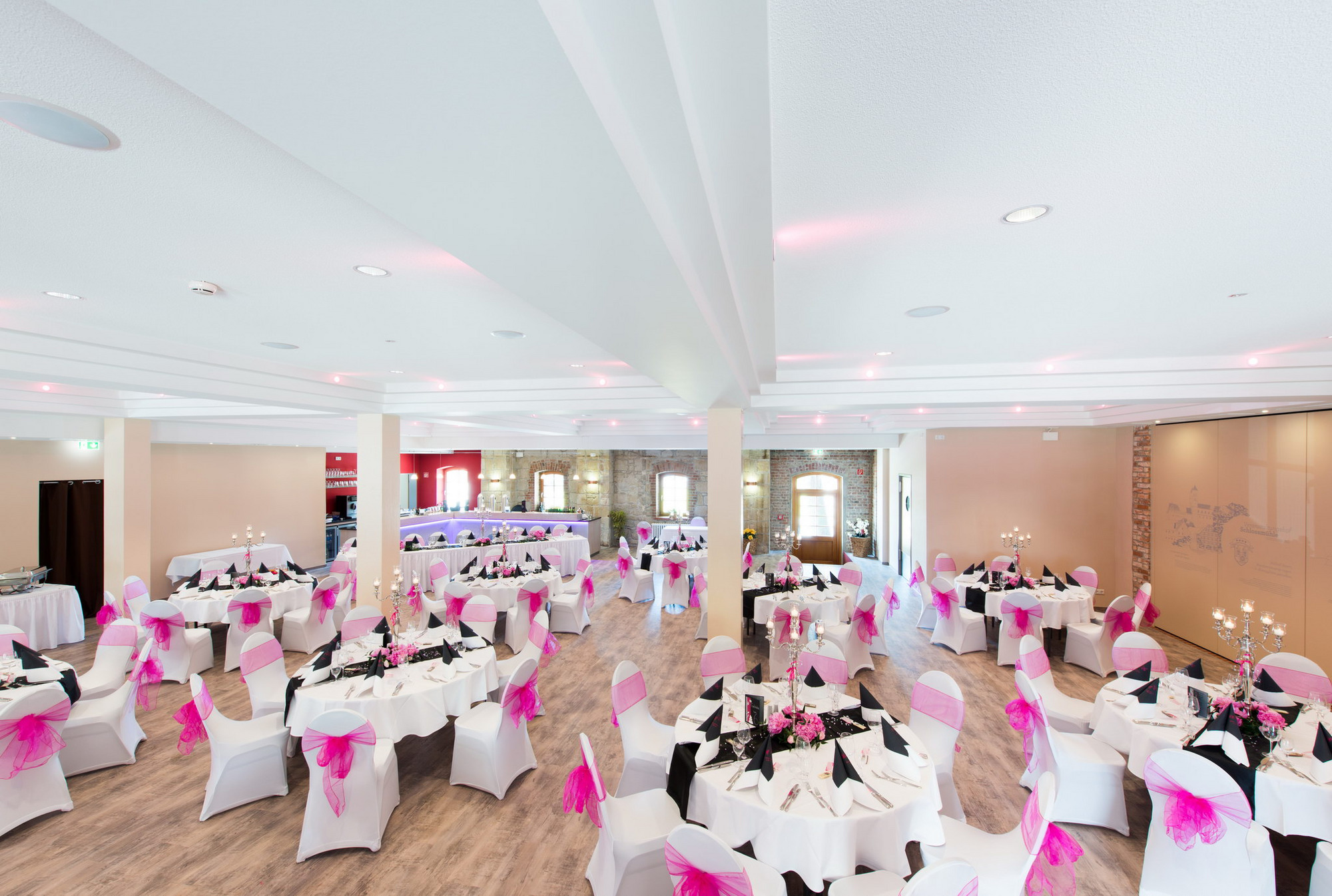 Eleganter Festsaal mit runden Tischen, pinken Blumen, weißen Hussen und Candelabern, im Hintergrund eine Bar.