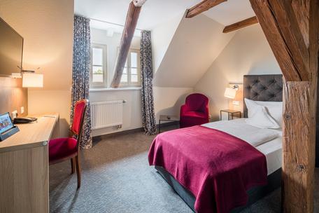 Gemütliches Schlafzimmer mit Dachschräge, Bett mit roter Decke, Schreibtisch, Sessel und zwei Fenstern.